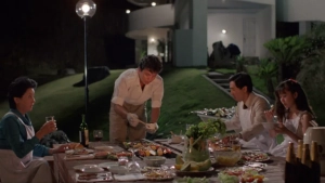 A group of four people dine at a long table filled with food and drinks outdoors at night, set against a modern white building and lush greenery.