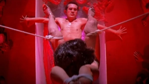 Angel Guts Nami 1979 red room scene. Nude man suspended in a glass tube, legs spread and bound by ropes while spectators watch the display.