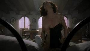 Anna Galiena standing topless in a black lingerie top with one breast exposed in a dimly lit bedroom scene.
