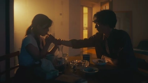 A cinematic shot of Remi Miyamori and her partner sharing an intimate moment over a small table in a dimly lit room from Like Love (Ai no Gotoku).