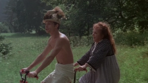 Park bench scene from Private Vices Public Pleasures (1976) - One individual wears a tall conical hat and feathered partial nude attire while conversing with another in white robes; horse-drawn carriage and riders appear in the forested background. - img #9