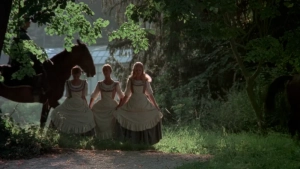 Park bench scene from Private Vices Public Pleasures (1976) - One individual wears a tall conical hat and feathered partial nude attire while conversing with another in white robes; horse-drawn carriage and riders appear in the forested background. - img #7