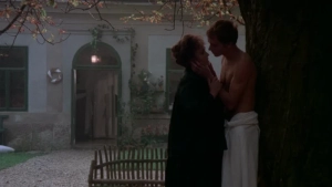 Park bench scene from Private Vices Public Pleasures (1976) - One individual wears a tall conical hat and feathered partial nude attire while conversing with another in white robes; horse-drawn carriage and riders appear in the forested background. - img #14