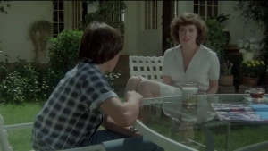 Close-up of actress Sylvia Kristel in an intimate conversation scene from *Private Lessons* (1981), wearing her signature short, curly hairstyle. She is seated outdoors at a glass patio table, engaged in a focused discussion with the young man across from her.