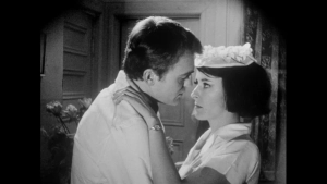 Black-and-white close-up from vintage erotic film Seeds of Sin (1968) showing a man and woman in an intense intimate embrace. The actress wears a classic maid's uniform with a frilly headpiece as she gazes deeply into her partner's eyes during this domestic scene, captured within a round-letterbox frame.