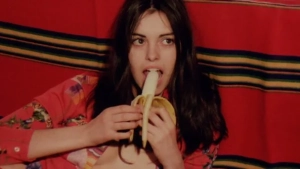 Lina Romay in open floral blouse eating a banana - intimate close-up shot against a red striped background from The Hot Nights of Linda (1975).