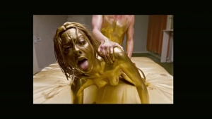Topless and nude intimate scene - woman and man covered entirely in shimmering gold body paint, kneeling on a bed.