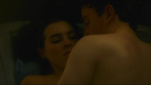 Topless intimate close-up in bed - Safiya Benaddi and shirtless partner cuddling or sleeping in a dimly lit scene from Truly Naked (2026).