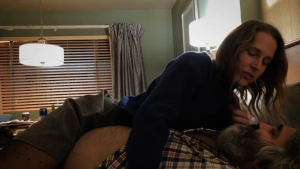 Josephine Decker leans over a shirtless older man lying on a bed in a hotel room, wearing a dark blue sweater and shorts pulled down slightly as she looks down at him closely.