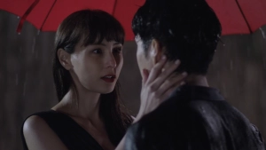 Lee Yu-jin standing under a red umbrella during a rain scene, gazing closely at a man whose back is turned to the camera as she gently touches his jaw with her hand.