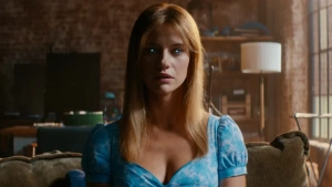 A medium shot of actress Lily Sullivan sitting on a sofa wearing a low-cut blue floral dress and looking intensely at the camera in a sunlit apartment interior.